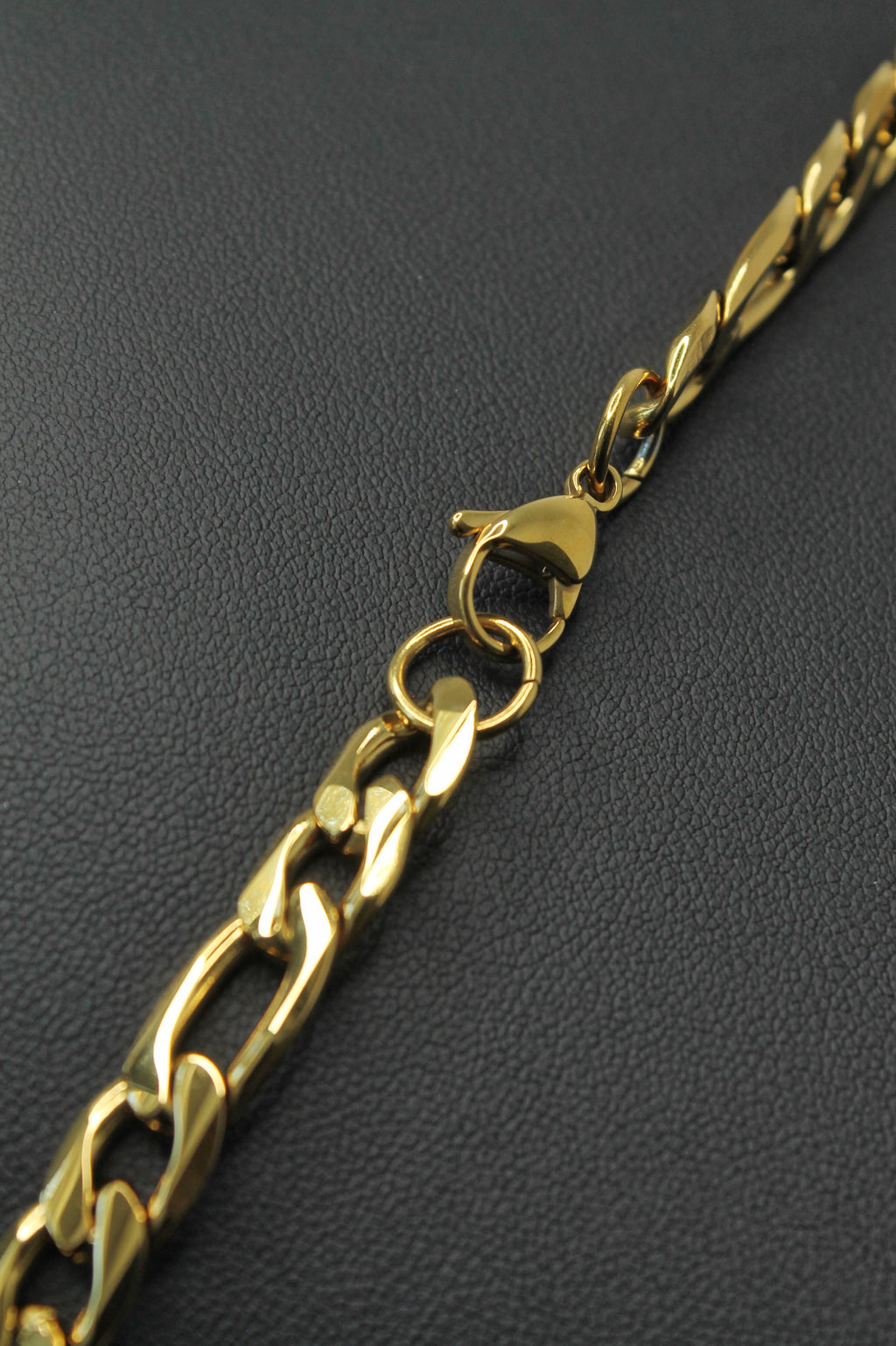 A close-up of a gold-tone Figaro chain necklace with a lobster clasp, laid diagonally on a dark textured surface.