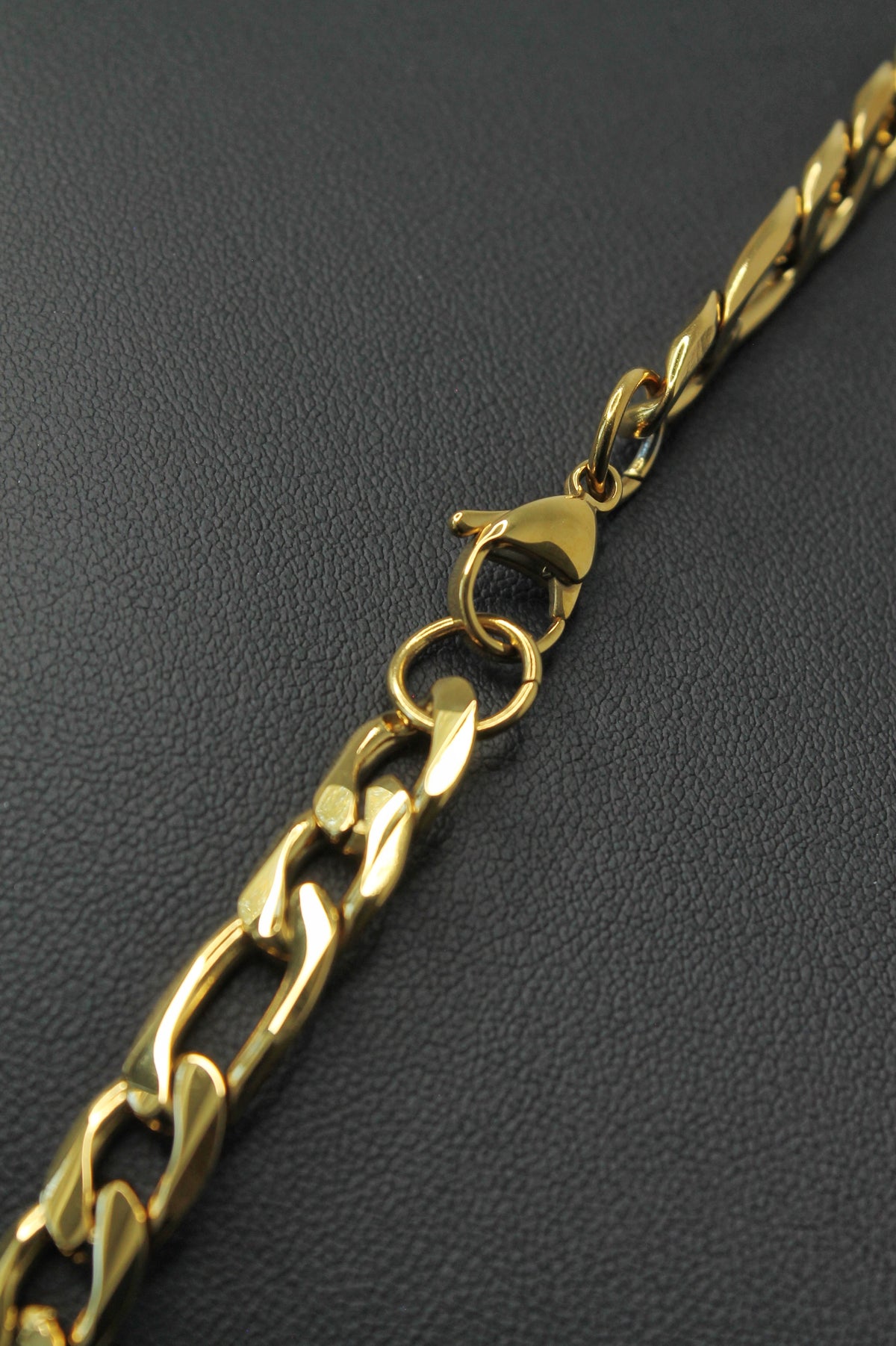 A close-up of a gold-tone Figaro chain necklace with a lobster clasp, laid diagonally on a dark textured surface.