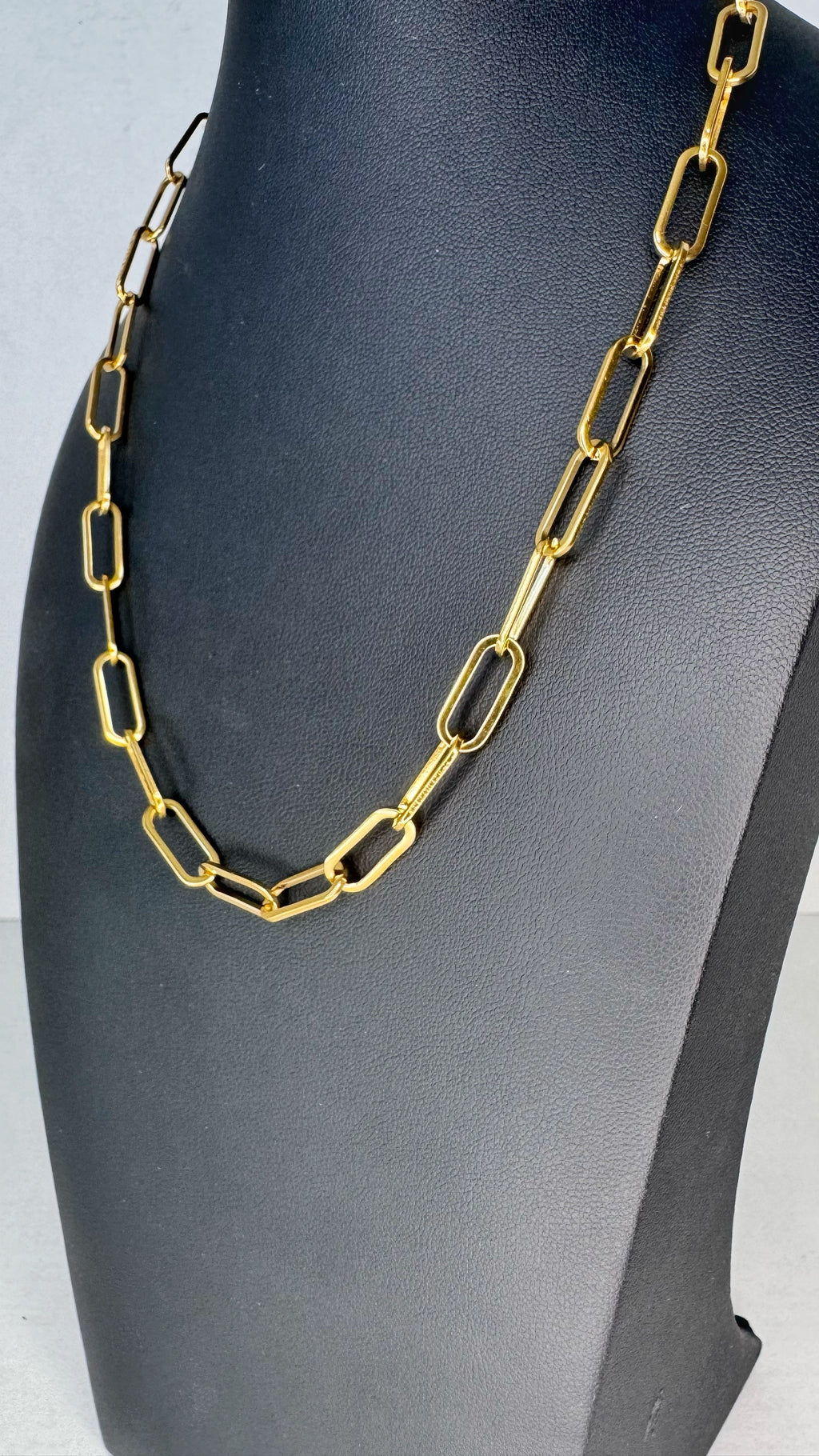 A gold paperclip chain necklace displayed on a black jewelry bust against a light gray background.