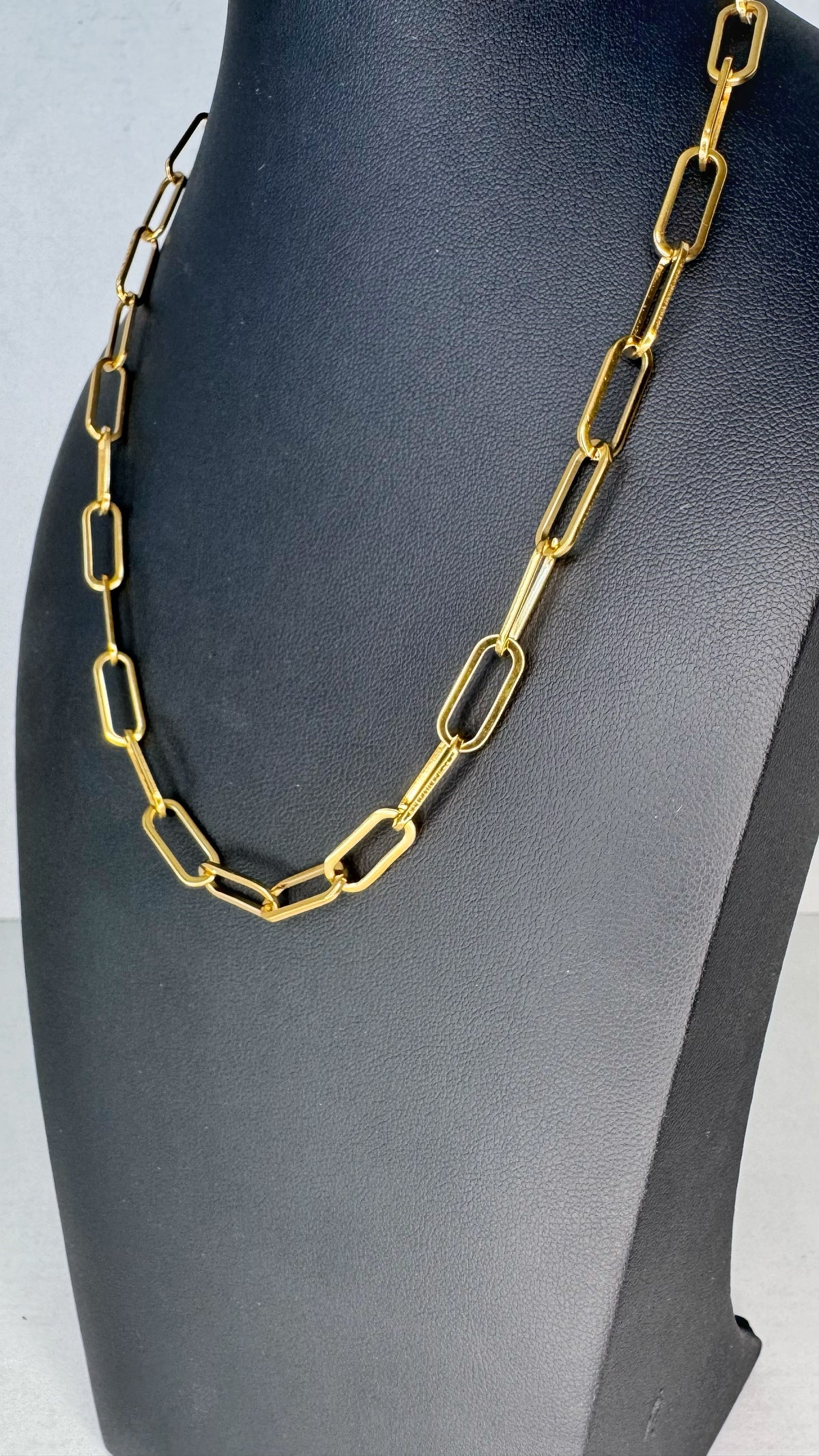 A gold paperclip chain necklace displayed on a black jewelry bust against a light gray background.