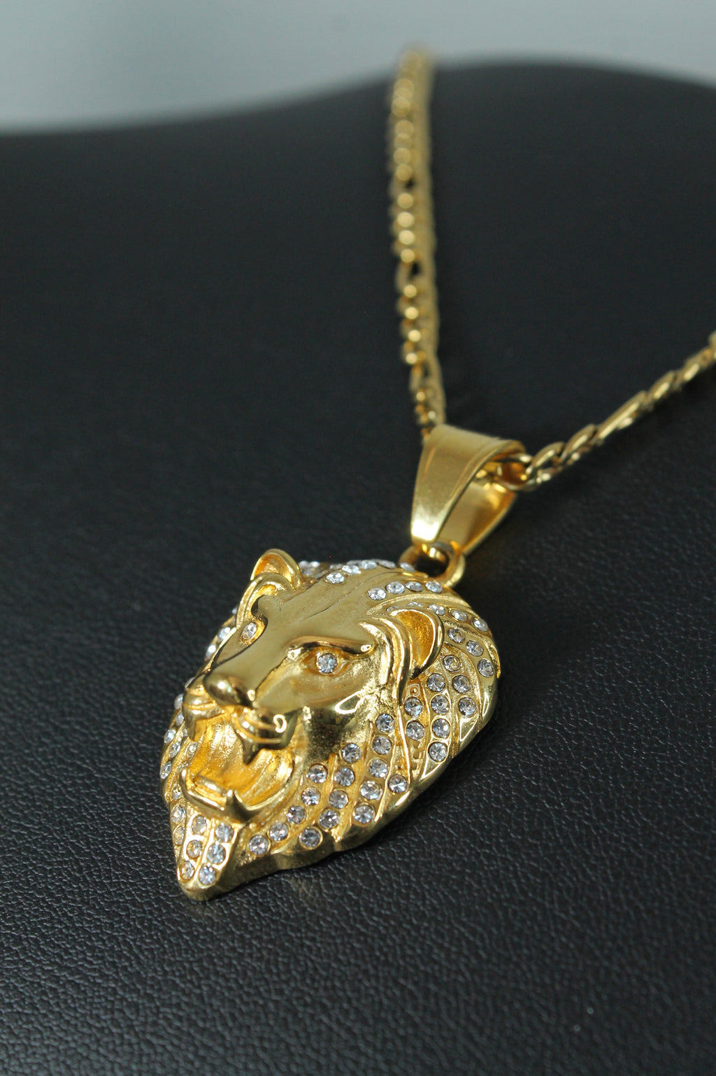 Gold lion head pendant necklace paved with small clear stones, displayed on a black textured surface.