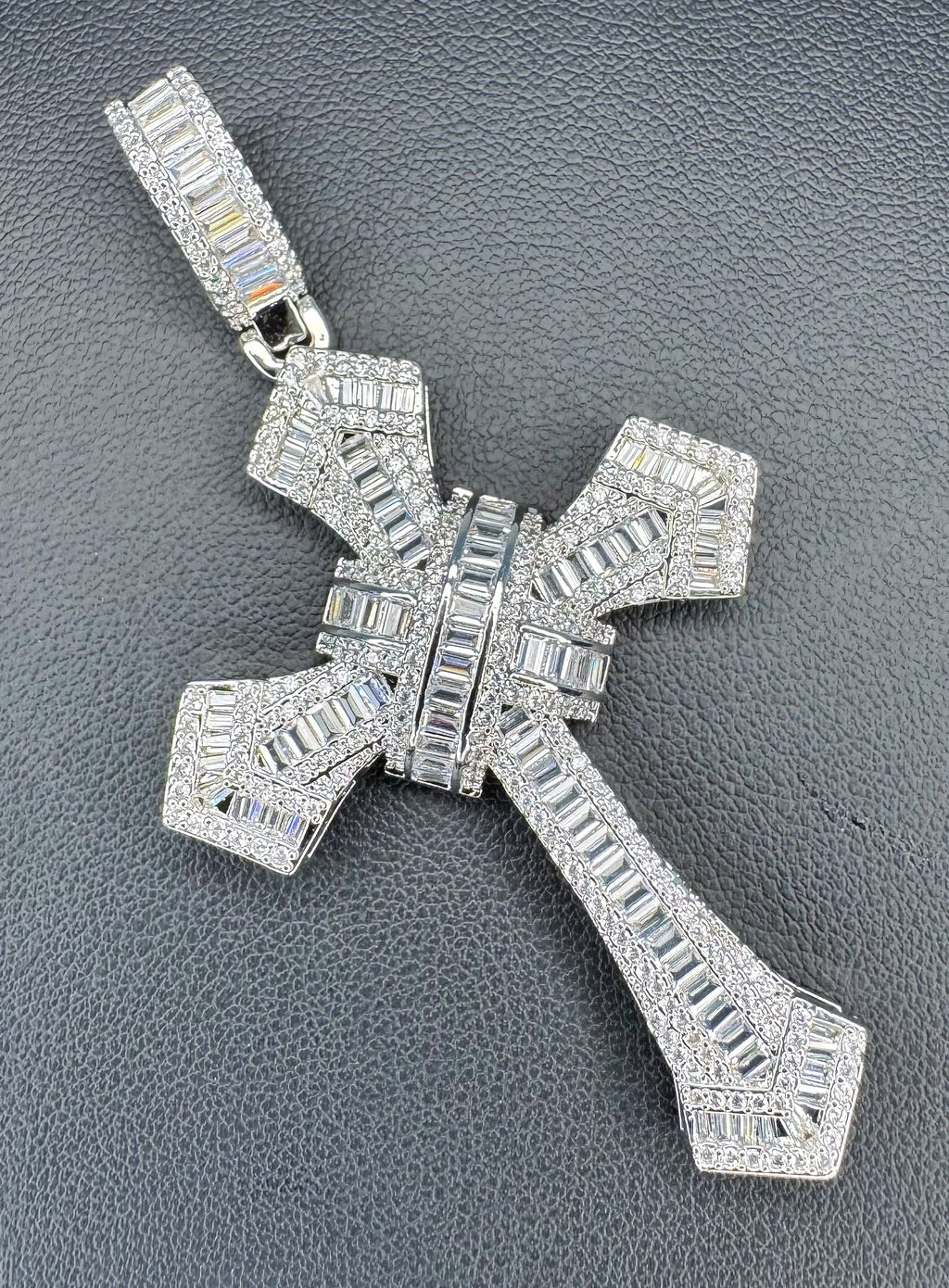 A silver cross pendant encrusted with clear baguette and round stones, displayed on a textured dark gray background.