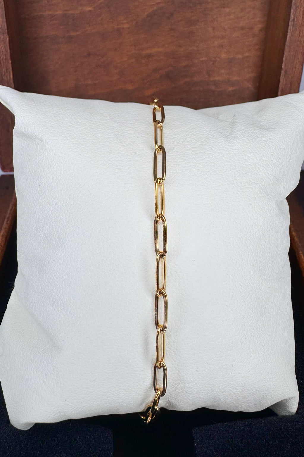 A gold paperclip chain link bracelet draped vertically down the center of a white textured pillow, set against a dark brown wooden background.