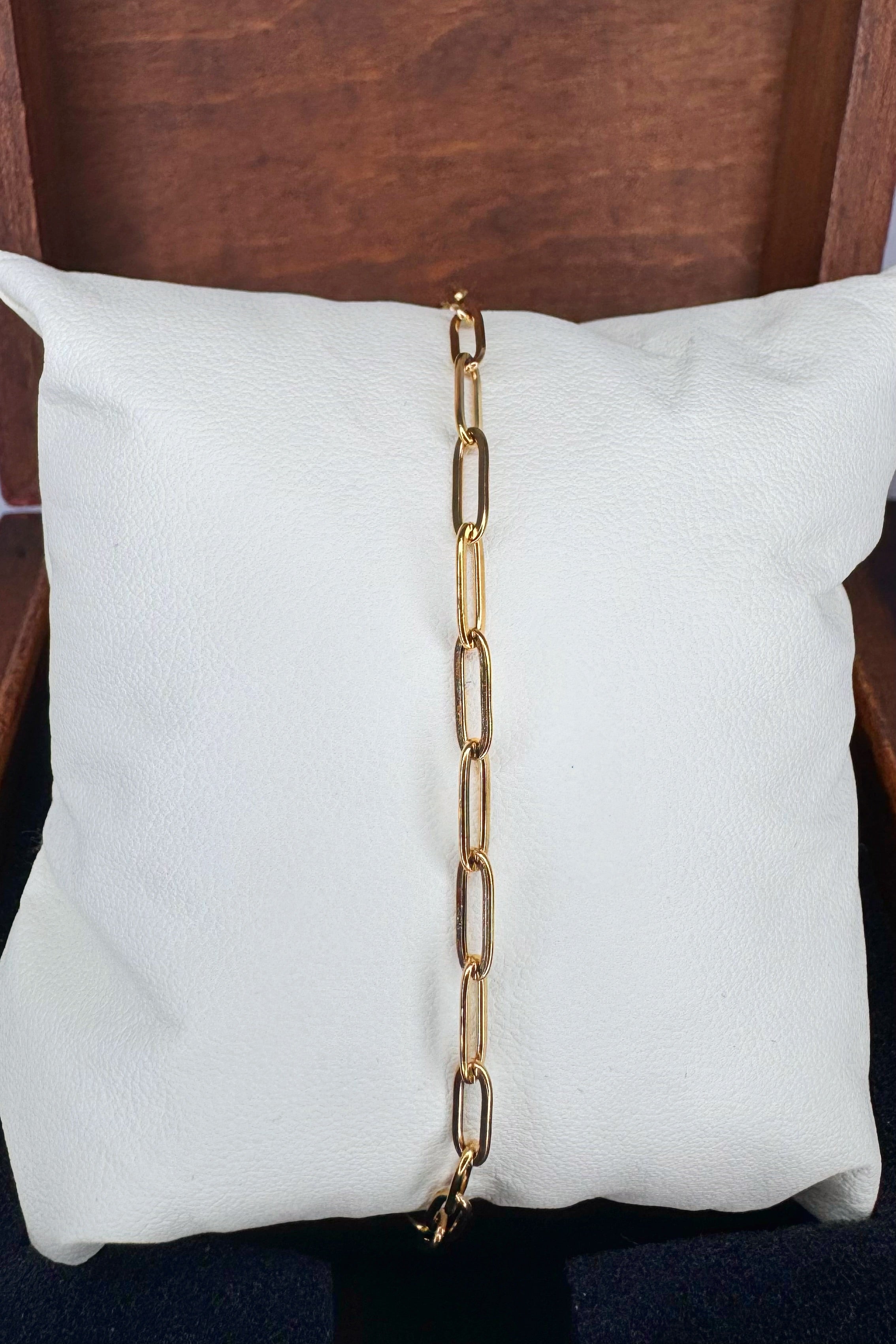 A gold paperclip chain link bracelet draped vertically down the center of a white textured pillow, set against a dark brown wooden background.