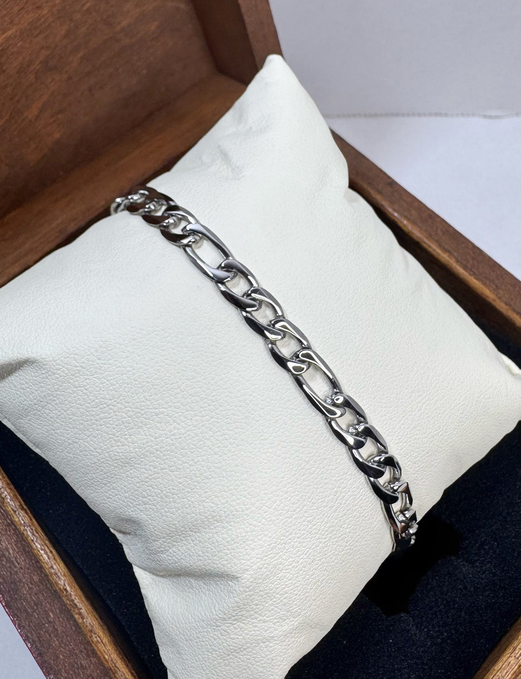 A silver-toned chunky figaro chain link bracelet displayed on a white cushion inside a wooden presentation box.