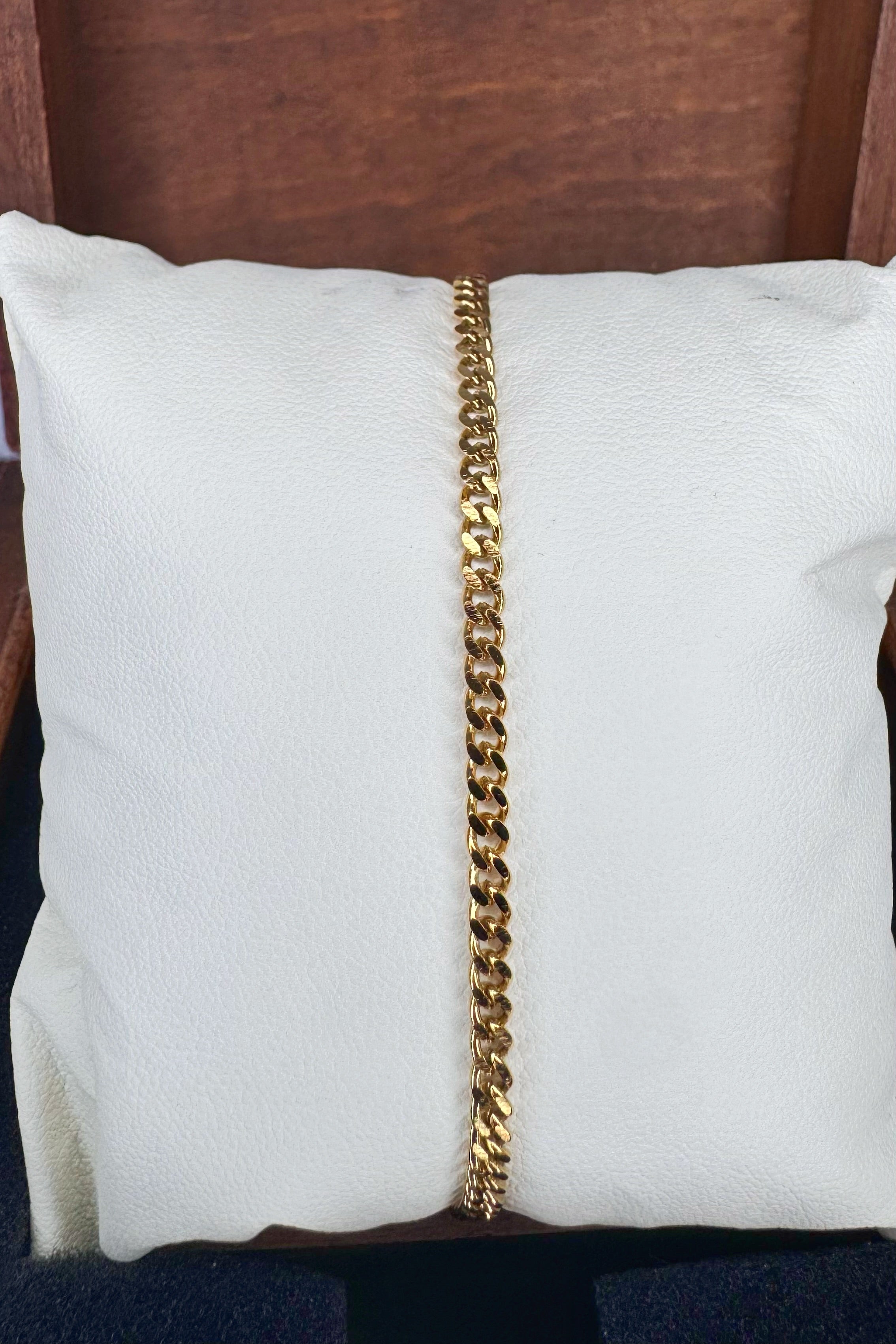 A gold Curb link chain bracelet displayed vertically on a white textured pillow, set against a dark brown wooden background.