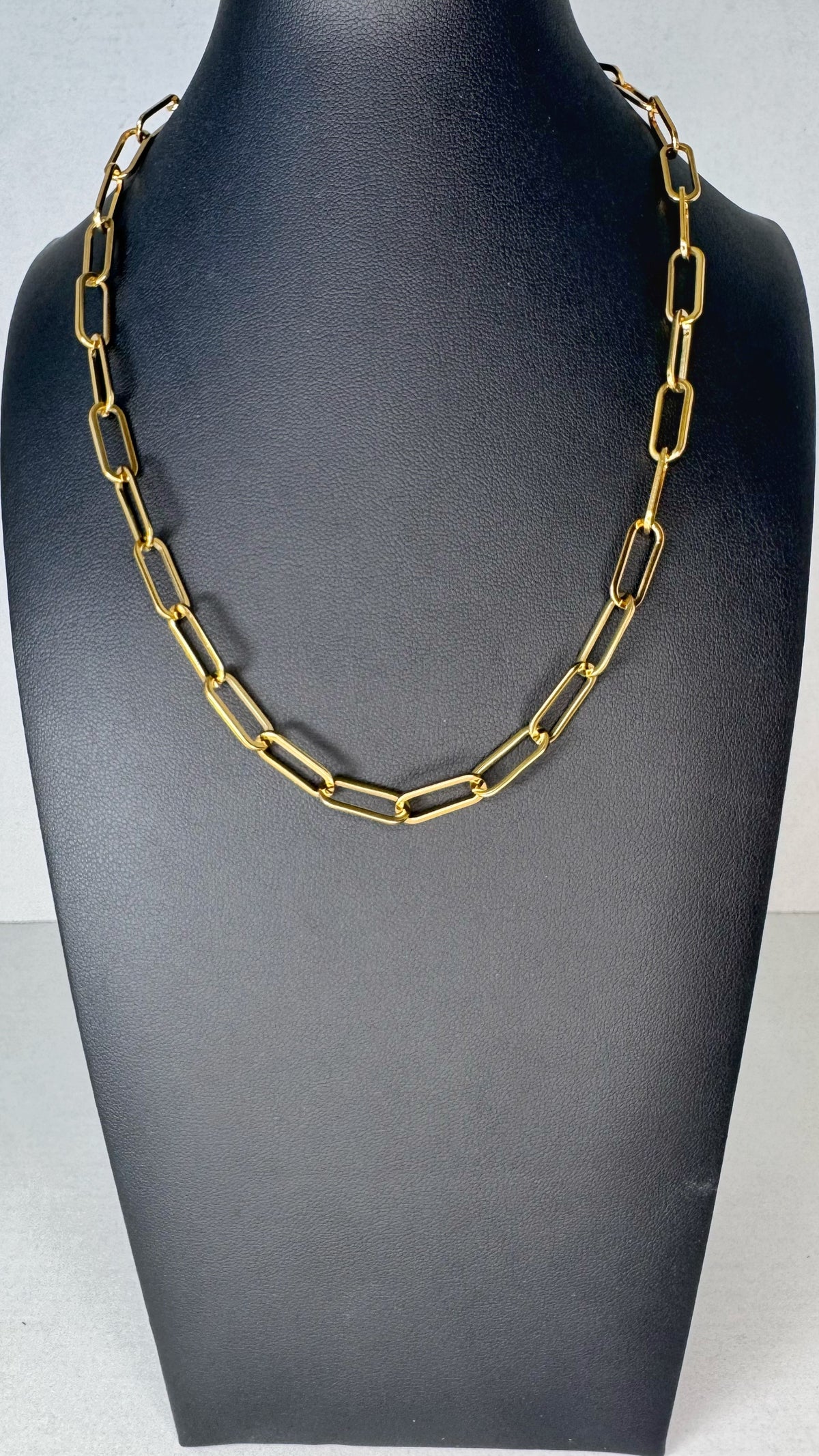 A gold-toned chain necklace with elongated rectangular links, displayed on a matte black jewelry bust against a light gray background.