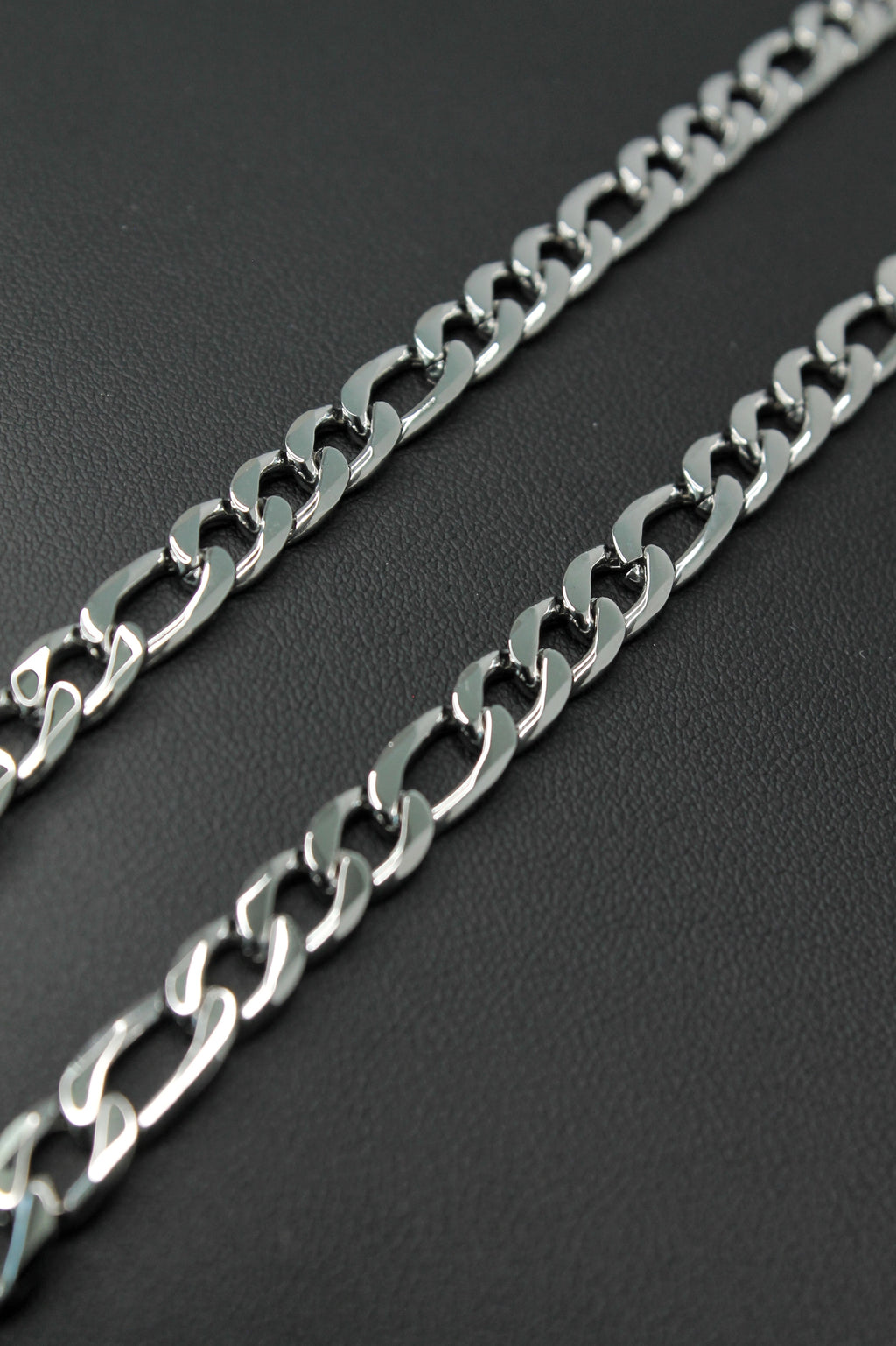 A close-up of a silver-toned metal Figaro chain laid diagonally across a dark textured background.