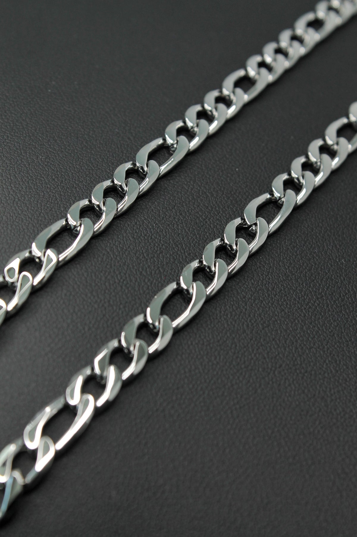 A close-up of a silver-toned metal Figaro chain laid diagonally across a dark textured background.