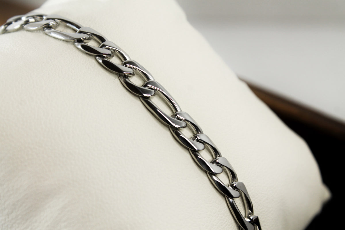 A close-up of a polished silver-toned metal figaro chain bracelet draped diagonally across a cream-colored display cushion.