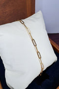 A gold paperclip chain bracelet draped over a white display pillow, set against a dark wooden chair and a light gray background.