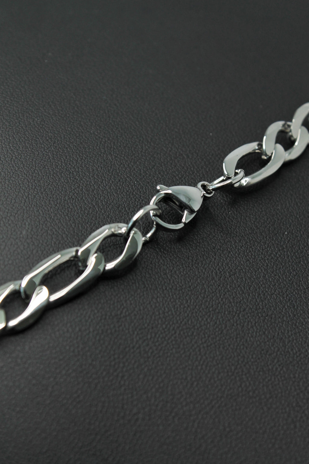 A close-up of a polished silver-tone metal Figaro chain with a lobster clasp, laid diagonally on a dark textured surface.