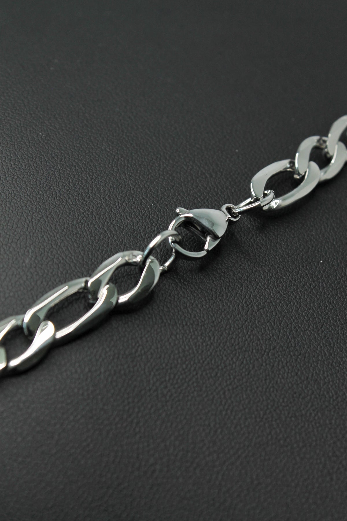 A close-up of a polished silver-tone metal Figaro chain with a lobster clasp, laid diagonally on a dark textured surface.