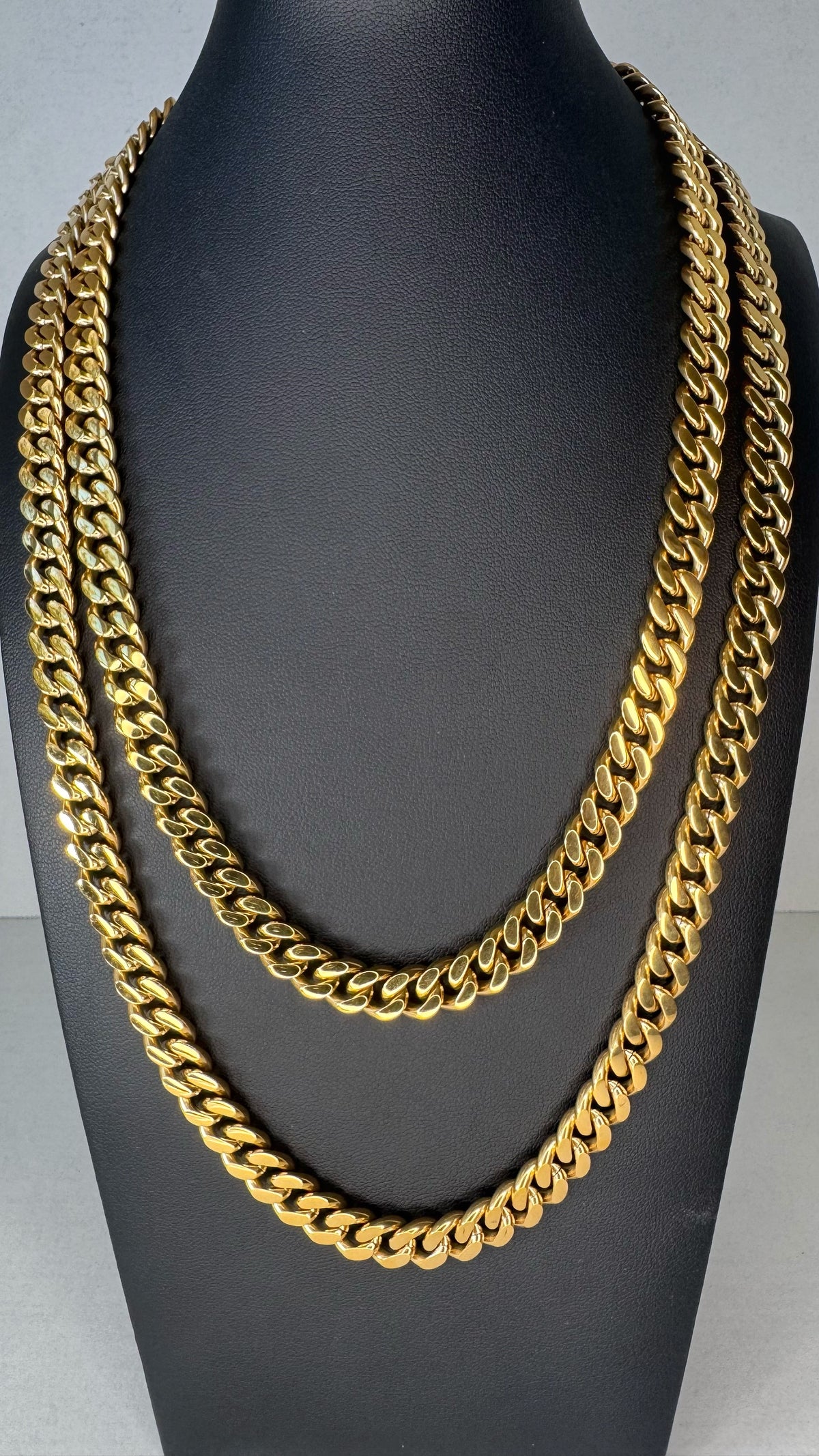 Two thick gold Curb link chain necklaces displayed on a black mannequin bust against a light gray background.